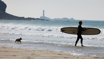 Surfer and dog Longships beach This landscape photograph captures a surfer carrying a longboard along the shoreline of Longships beach, with a dog running at the water’s edge, during a spring afternoon. The scene is set on the coast of Sennen Cove in Cornwall, United Kingdom, where gentle waves break on the sandy beach. In the distance, the Longships Lighthouse is visible on its rocky outcrop, providing a recognizable landmark along the rugged coastline. The image highlights the natural environment of this popular beach destination, focusing on the activity of surfers enjoying the outdoors amid the scenic beauty of Cornwall’s coastline.